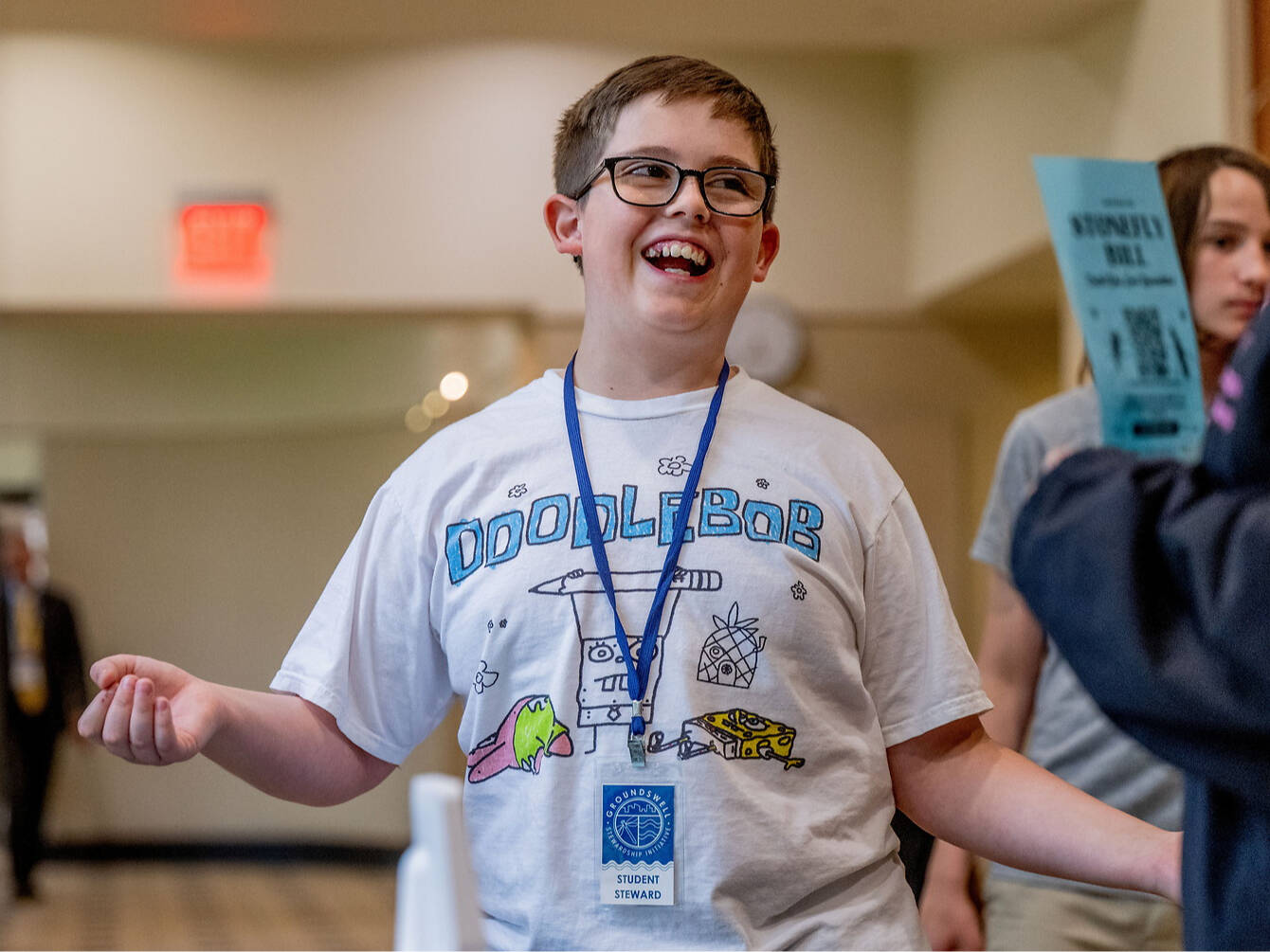 A smiling k-12 student wearing glasses and a "DoodleBob" t-shirt with a student steward badge stands in a hallway, gesturing with their hands.